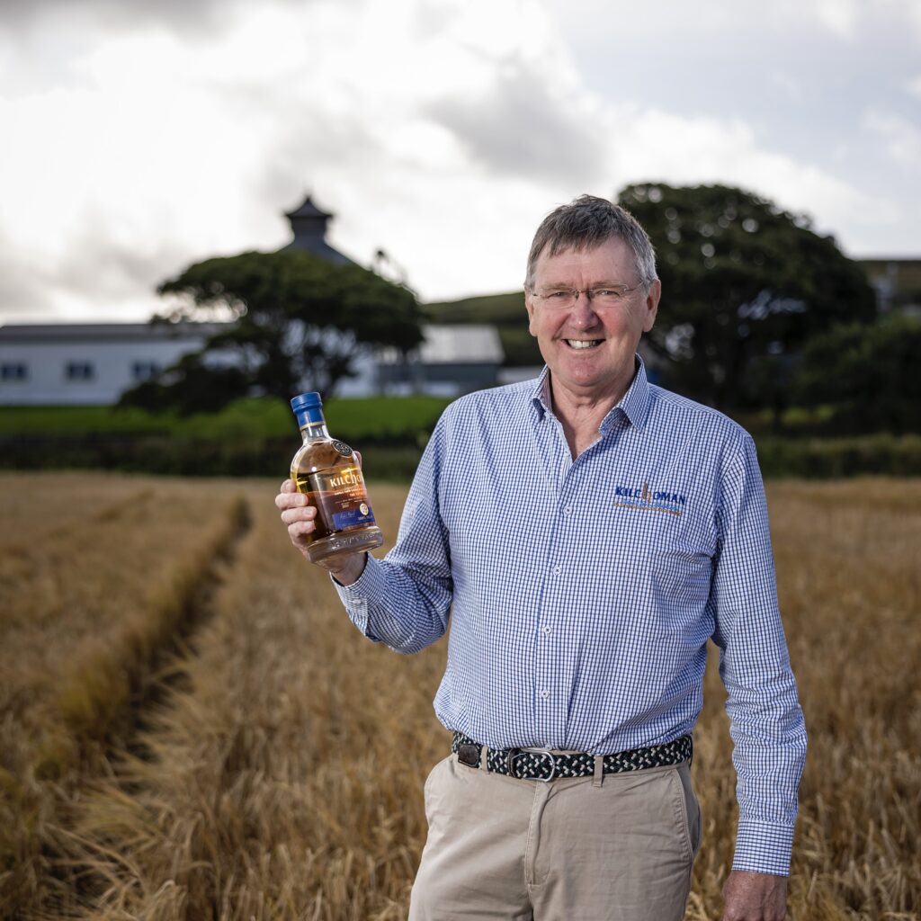 Anthony Wills Kilchoman barley field holding a bottle of 100% Islay single malt scotch whisky